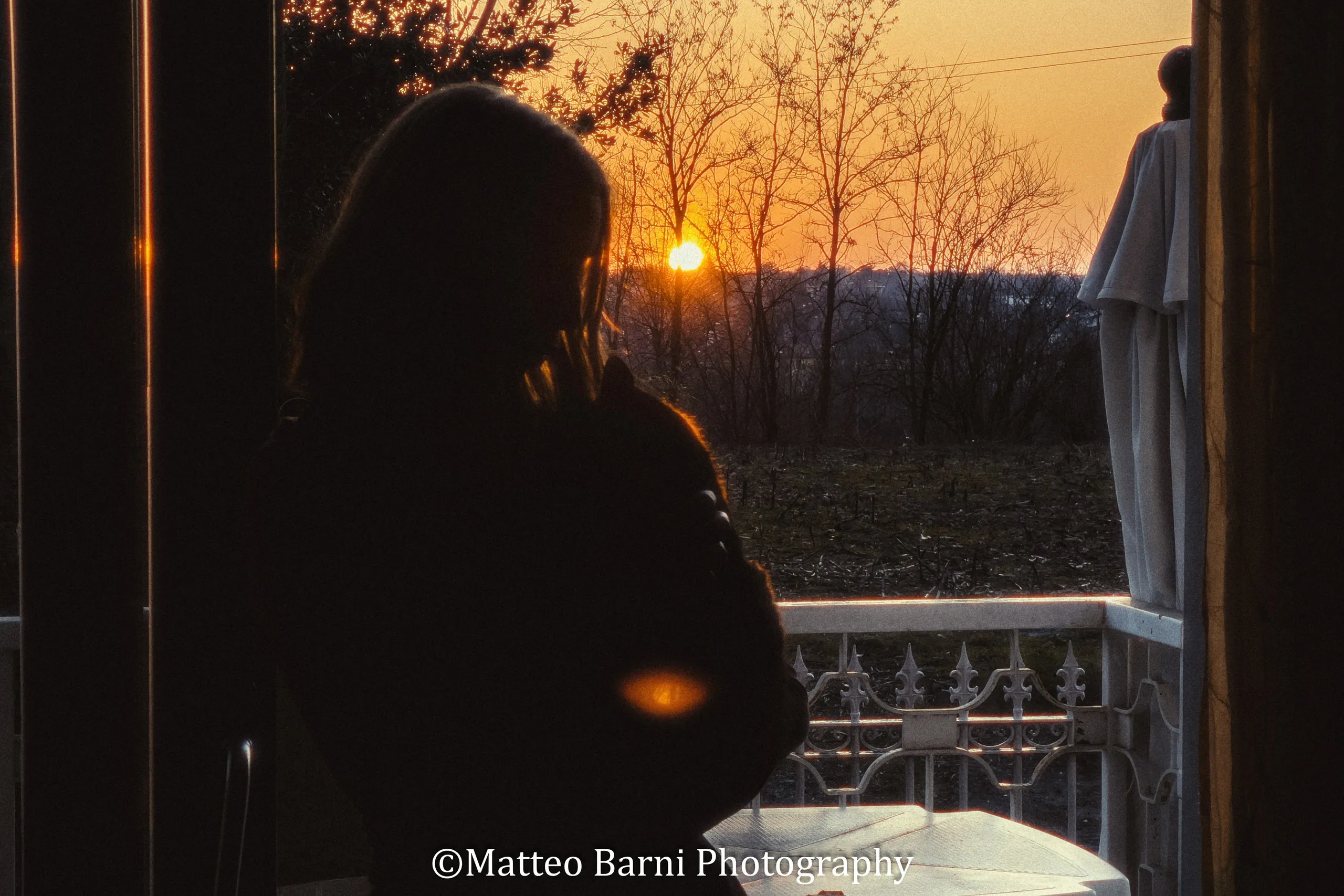 Una ragazza in controluce sulla porta finestra di un balcone, con il sole che tramonta dietro gli alberi