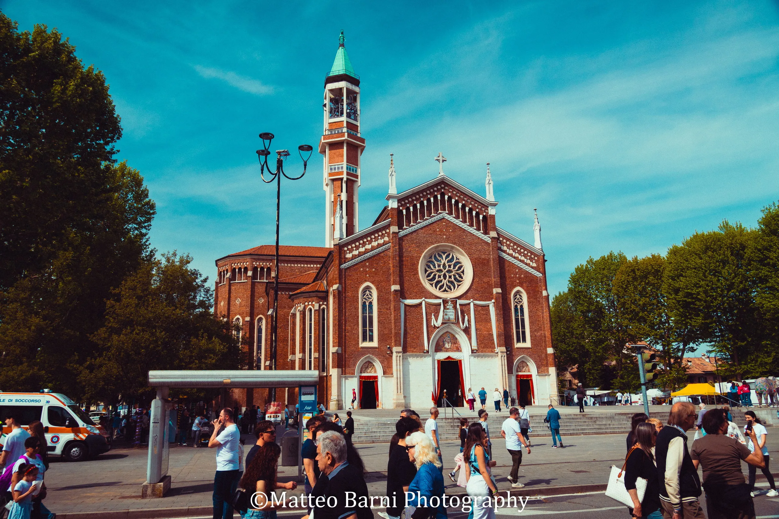 Folla nella piazza davanti a una chiesa di mattoni rossi con il campanile, sotto un cielo azzurro