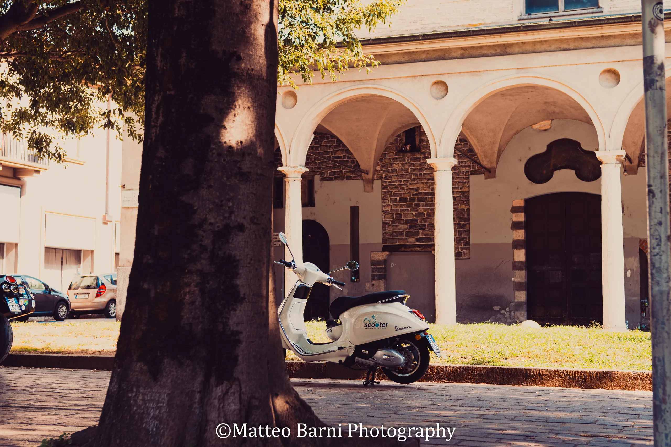 Uno scooter bianco parcheggiato sotto un portico ad archi, accanto al tronco di un albero